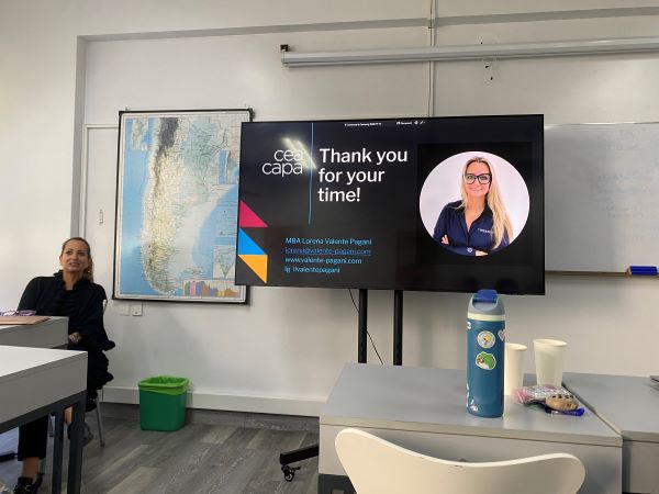 A person sitting at a desk at the front of a classroom next to a TV screen with a slide reading