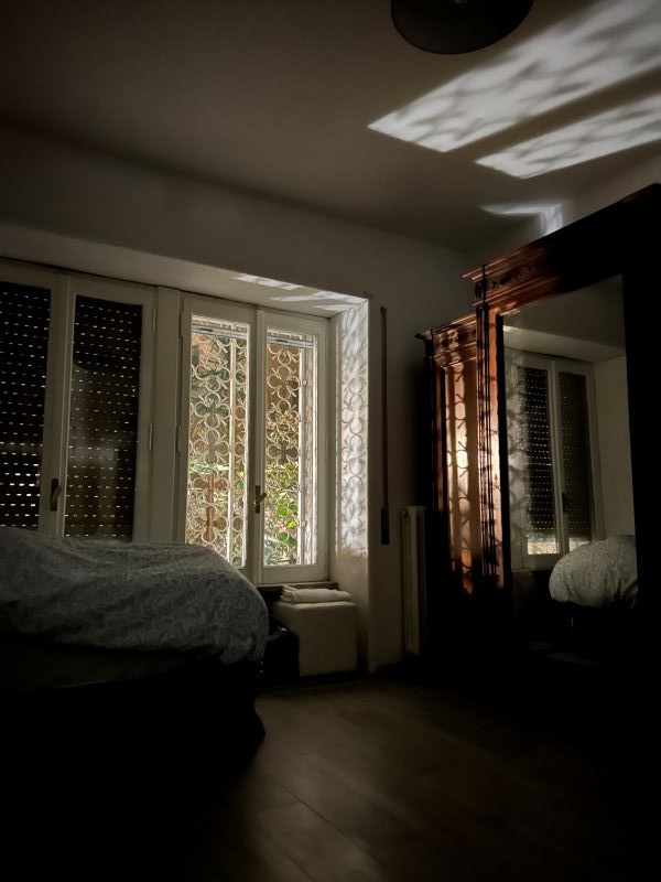 A bedroom seen in shadowy light, featuring a large window in the middle of the frame letting in morning light