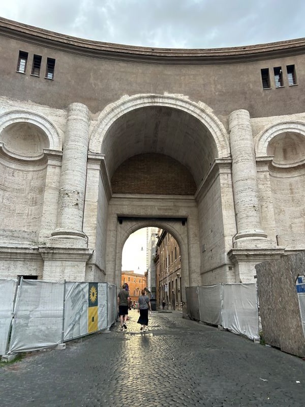 A cobblestone street underneath a large archway, featuring two people walking away from the camera underneath the archway