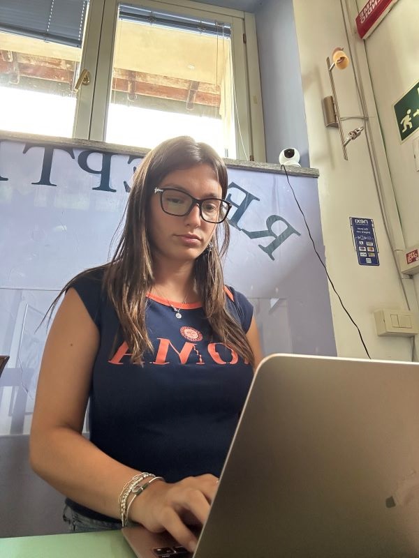 A study abroad student wearing glasses and standing at a desk indoors, looking down at the laptop they are typing on