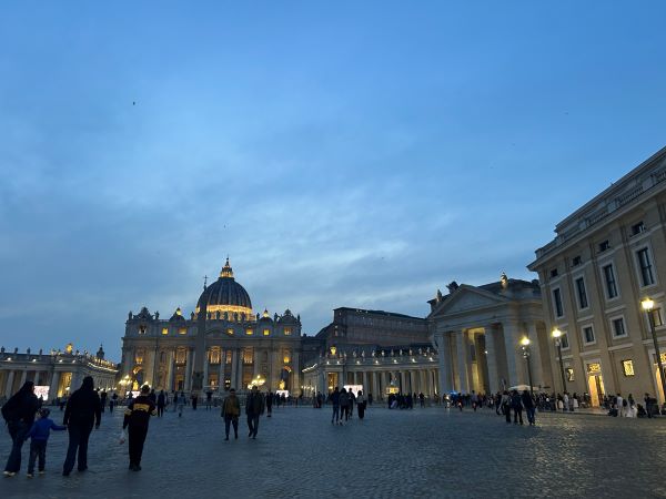 A large ornate building lit up with bright golden lights as people walk around the square in front of it, all under a dark blue evening sky
