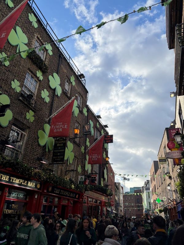 A city street full of people, under a cloudy blue sky and with buildings on either side decorated with large green shamrock signs