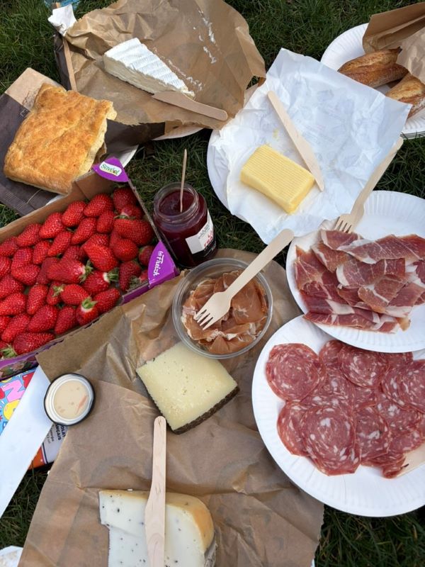 A picnic of food on paper and plates on top of grass, featuring sliced meats, chunks of cheese, butter, a jar of jam, and a box of bright red strawberries