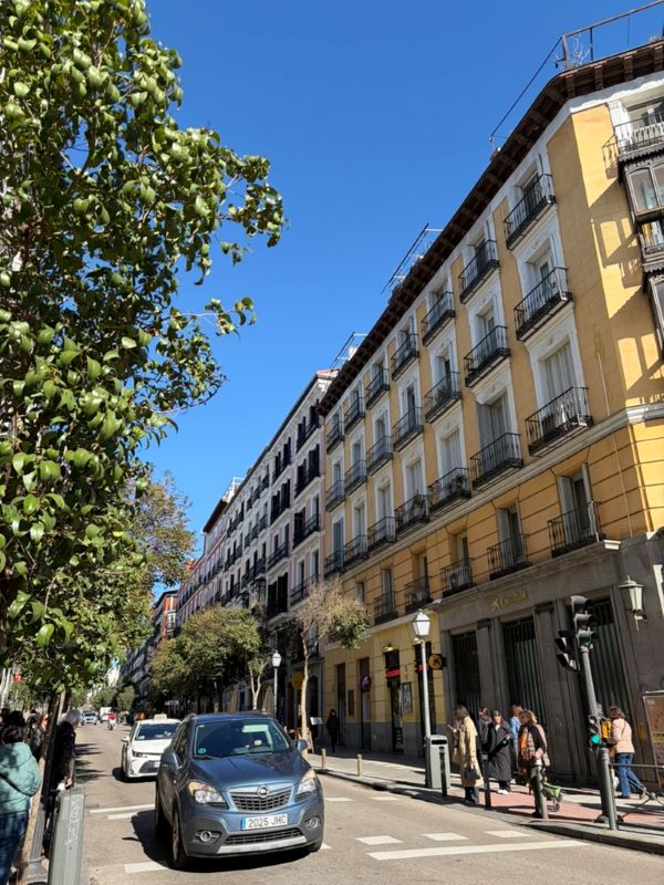 A city street with cars driving down it and a creamy yellow building on the righthand side on a bright sunny day