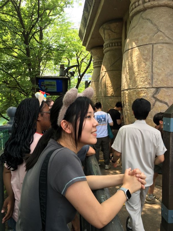 A study abroad student wearing a headband with small fake fluffy animal ears smiling and looking away from the camera while leaning on a railing outdoors