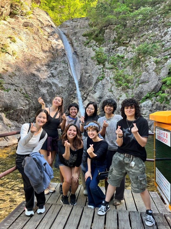 A group of study abroad students standing outdoors in front of a body of water and making small heart shapes with their fingers while smiling at the camera