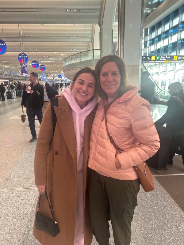 A study abroad student standing with her mother inside an airport, both smiling at the camera