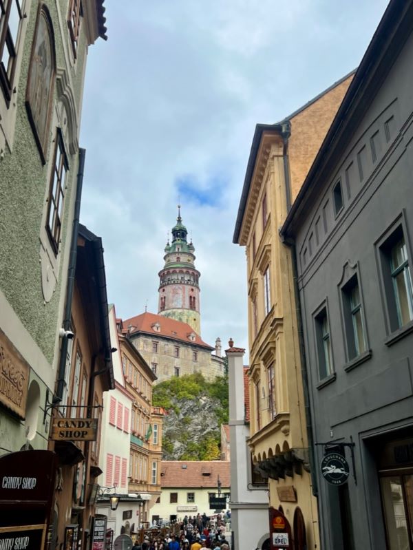 A large castle seen from the bottom of a street, with tall buildings on either side and many people on the street leading to the castle