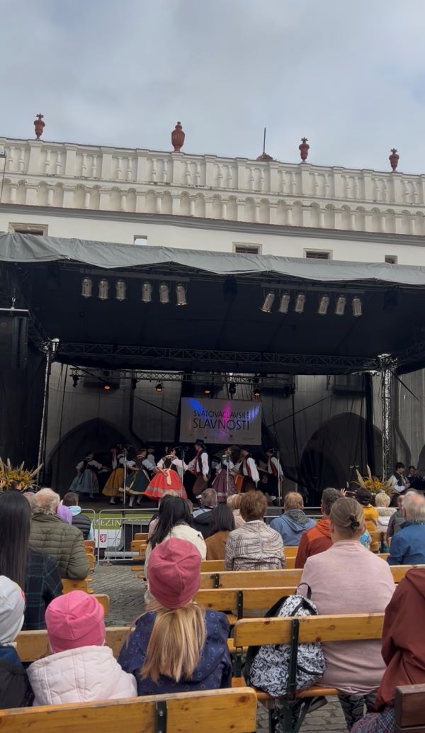 People sitting on rows of outdoor benches watching dancers perform on a covered outdoor stage