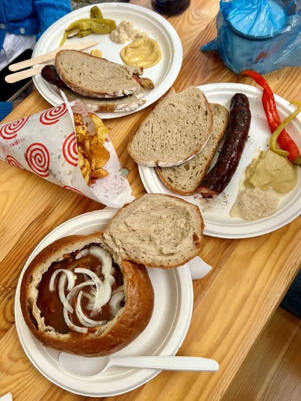 A table covered with plates of food, including bread, a sausage, soup with onion slices, and potato slices