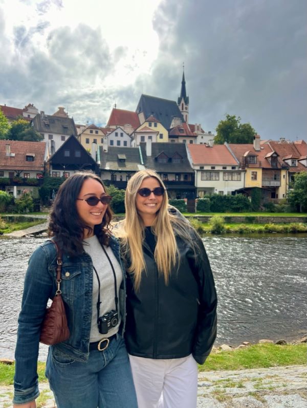 Two study abroad students wearing sunglasses, standing in front of a river and town buildings, smiling and looking slightly away from the camera