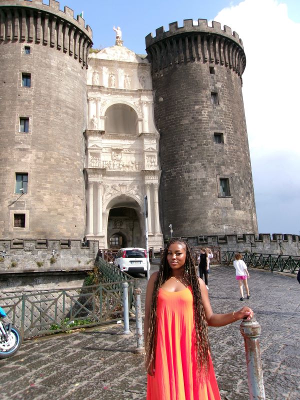 A study abroad student with long hair and wearing a bright orange dress standing outdoors in front of a tall ornate building and smiling at the camera