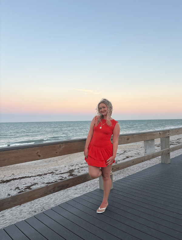 A study abroad student with curly blonde hair and wearing a red dress standing on a boardwalk in front of the ocean and smiling at the camera