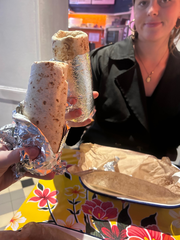 A study abroad student holding up a burrito to a hand from behind the camera also holding a burrito
