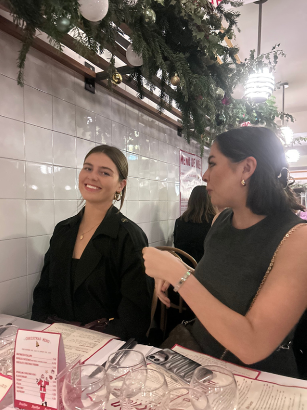Two study abroad students sitting an in indoor restaurant table, smiling while one looks at the camera and the other looks at their friend