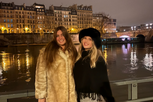 Two study abroad students standing outdoor in winter coats in front of a river at nighttime and smiling at the camera
