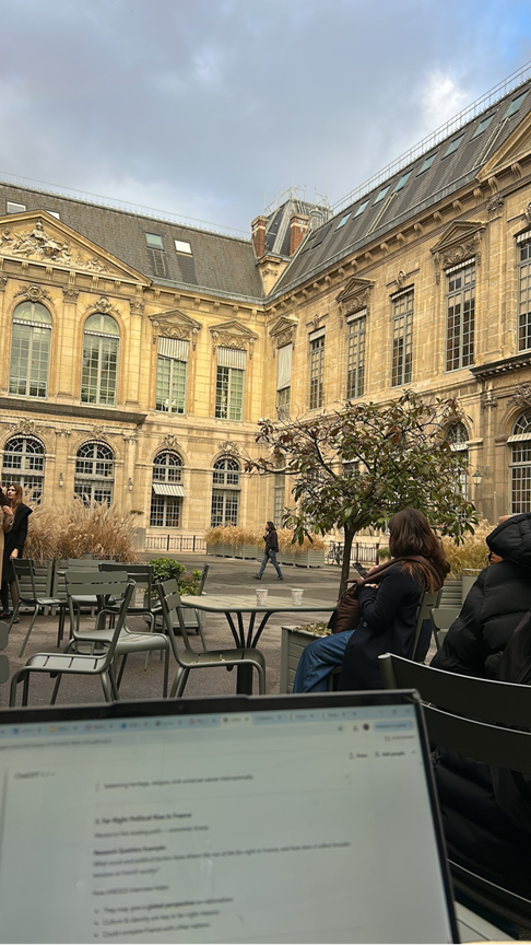 A blurry laptop screen seen in the foreground while in the background people sit at tables in an outdoor courtyard surrounded by tall ornate buildings