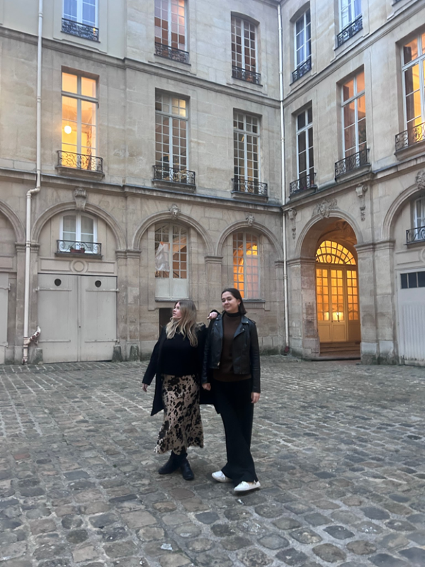 Two study abroad students standing outdoors on a cobblestone street in front of tall stone buildings, one smiling at the camera and the other smiling and looking away from the camera
