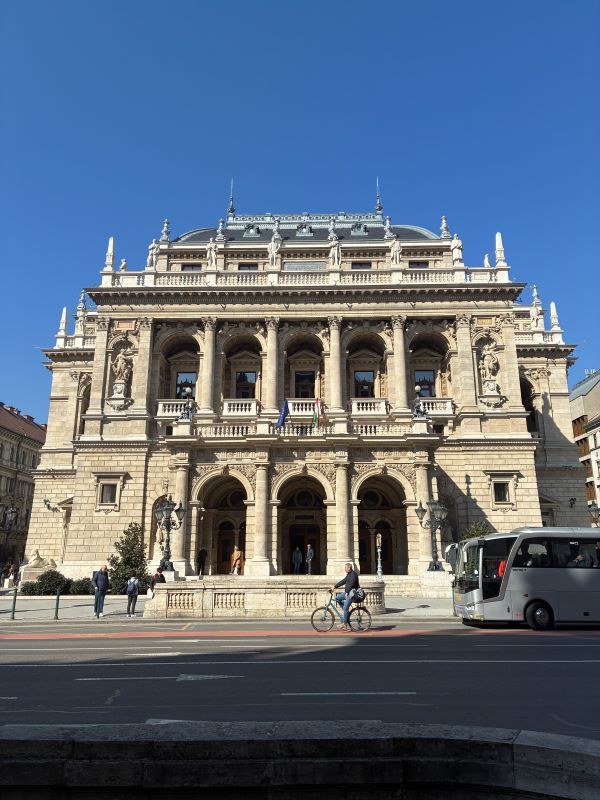 A large ornate sand-colored building with a busy road in the foreground and underneath a bright blue sky