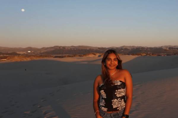 A study abroad student standing outdoors in front of a sandy landscape with the moon small in the distance, and smiling at the camera