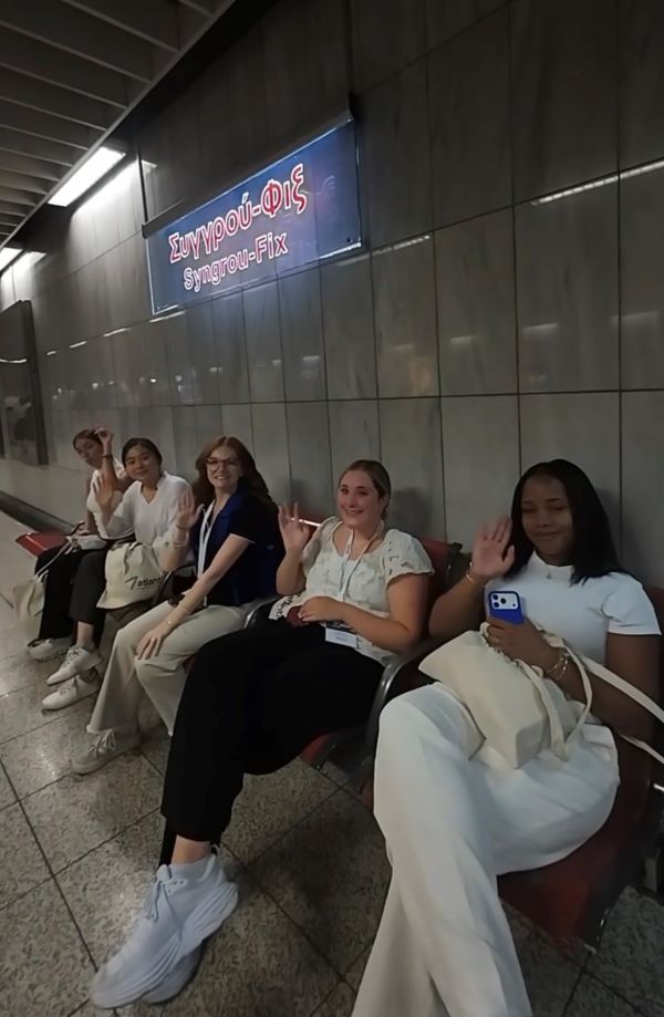 Five pre-med shadowing students sitting on a bench inside a metro station and waving and smiling at the camera