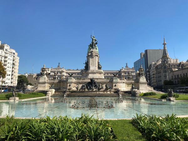 A large intricately designed building behind a small pond and surrounded by green grass under a bright blue sky