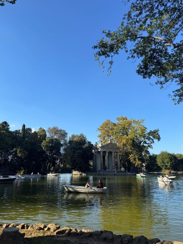 People sitting in small white rowboats in a pond seen mostly in shade provided by nearby trees with a small stone building in the background