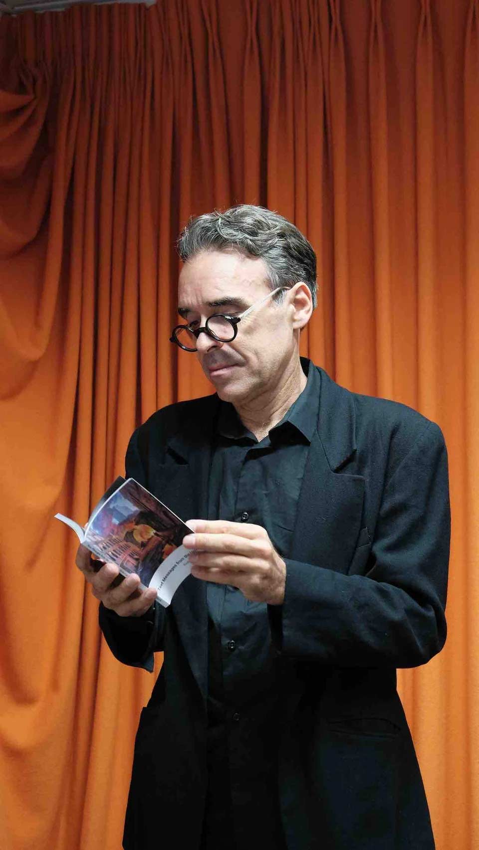 Dr. Richard James Allen standing up and reading a book while wearing glasses and a black shirt, blazer, and pants against a wooden wall background