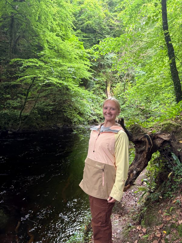 A study abroad student standing in front of a river in a wooded area and smiling at the camera