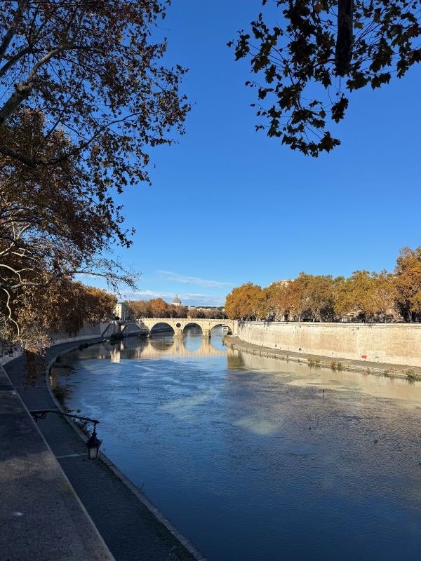 A river hemmed in by stone walls on either side, with a stone arched bridge seen intersecting the water further in the distance