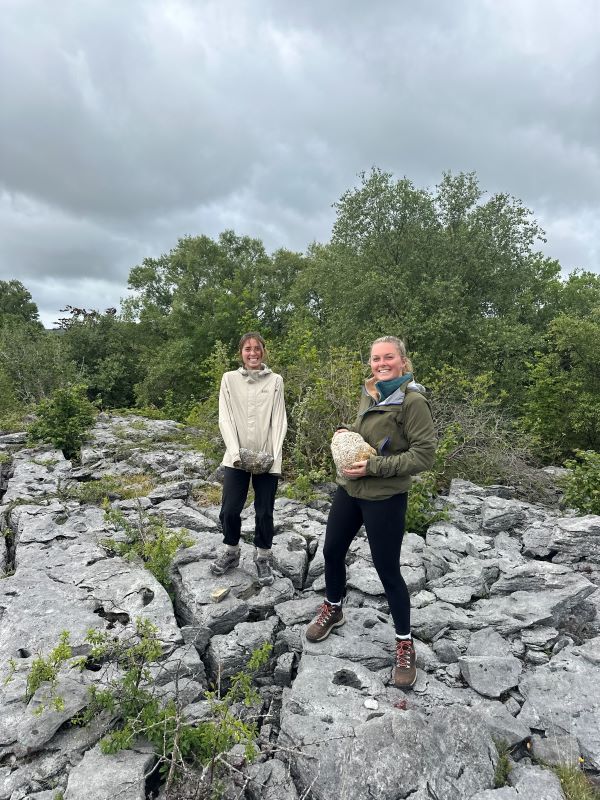 Two study abroad students standing on a rocky outdoor path and holding large rocks while smiling at the camera