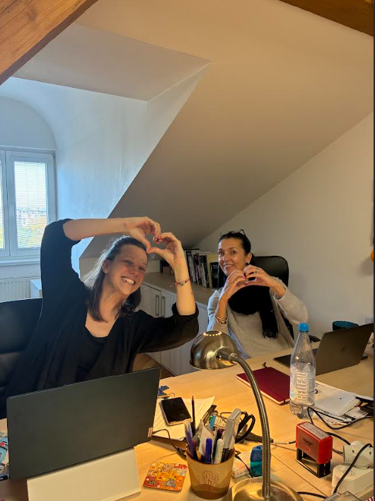 Two people sitting at a desk indoors and making heart shapes with their hands while smiling at the camera