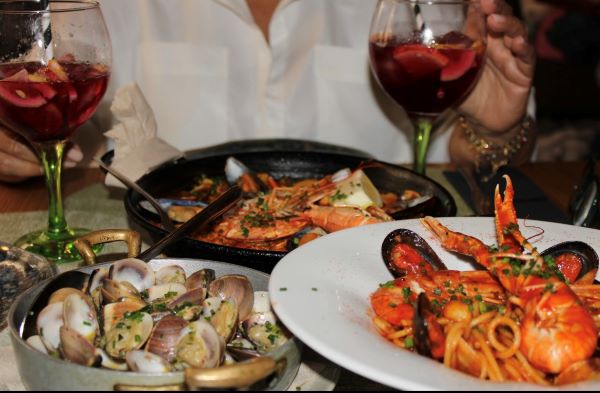 Colorful plates of seafood sat on a table alongside glasses of red sangria with a person seen from the head down sitting in the background
