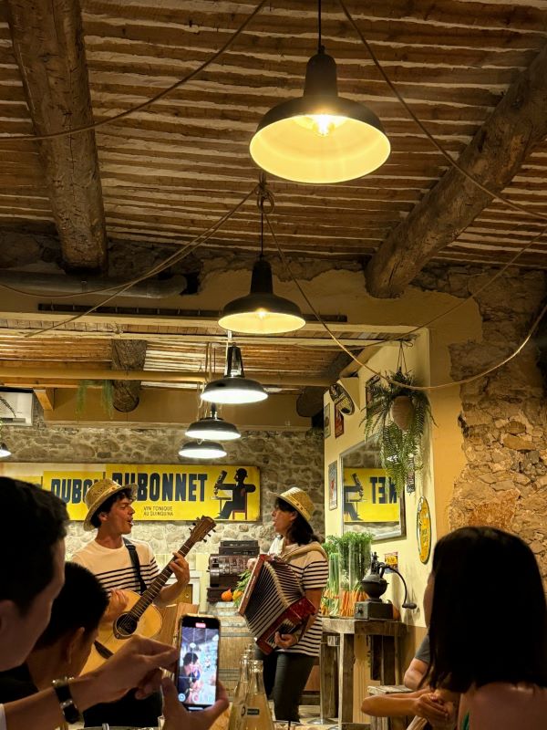 Two musicians wearing blue-and-white striped shirts and wearing small straw hats, one playing a guitar and one an accordion, performing in an indoor space