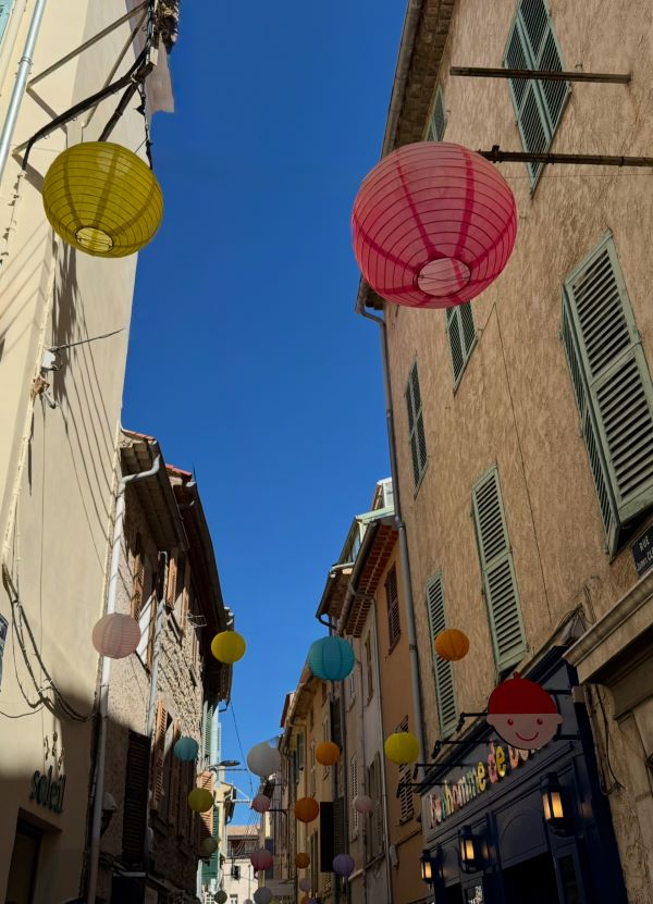 A narrow city street hemmed in by sand-colored buildings with brightly-colored paper lanterns hung between buildings and underneath a bright blue sky