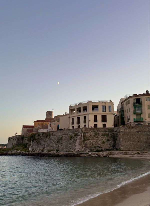 A collection of sand-colored buildings standing above water-level with a body of water in the foreground, seen in low evening light