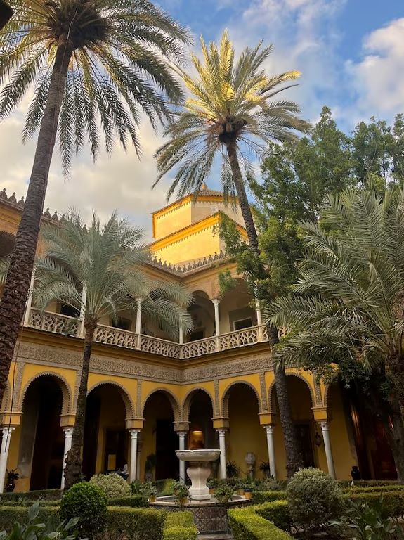 The exterior of a large intricately designed building with a small fountain and garden in the foreground and palm trees overhead