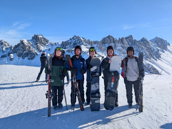 A group of study abroad students in winter sports gear holding skis and snowboards in a snowy outdoor setting