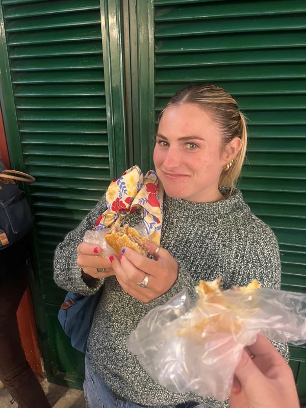 A study abroad student wearing a green sweater holding a half-eaten sandwich and smiling at the camera