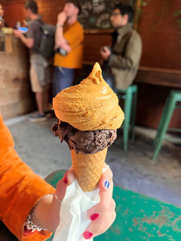 A hand with red-and-blue-painted nails holding an ice-cream cone with a two scoops of gelato, one dark brown and one a caramel color