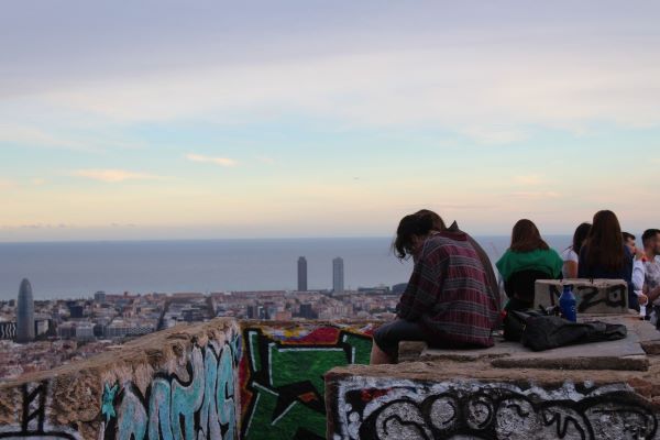 People sitting on low concrete walls with city buildings in the background underneath a sky full of soft colors