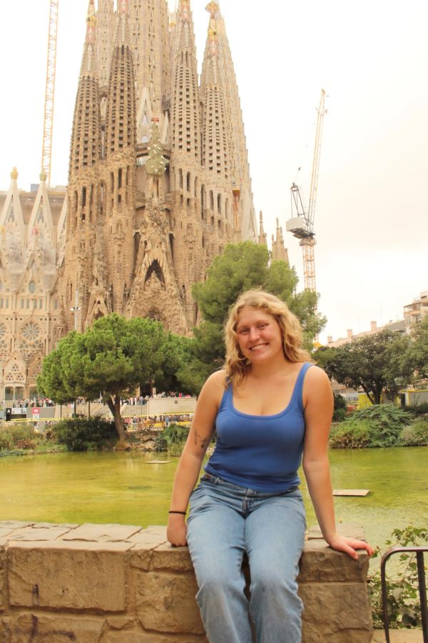 A study abroad student wearing a blue tank top and jeans smiling at the camera and sitting on a low stone wall in front of the massive Sagrada Familia church