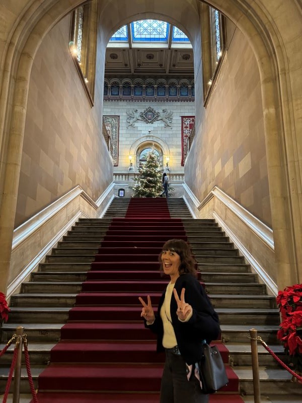 A study abroad professor standing at the bottom of a large indoor stone staircase, smiling at the camera and holding up their hands in peace signs