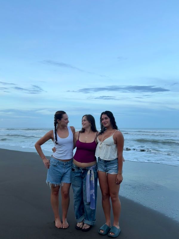 Three study abroad students standing on a beach in front of a large body of water in evening light