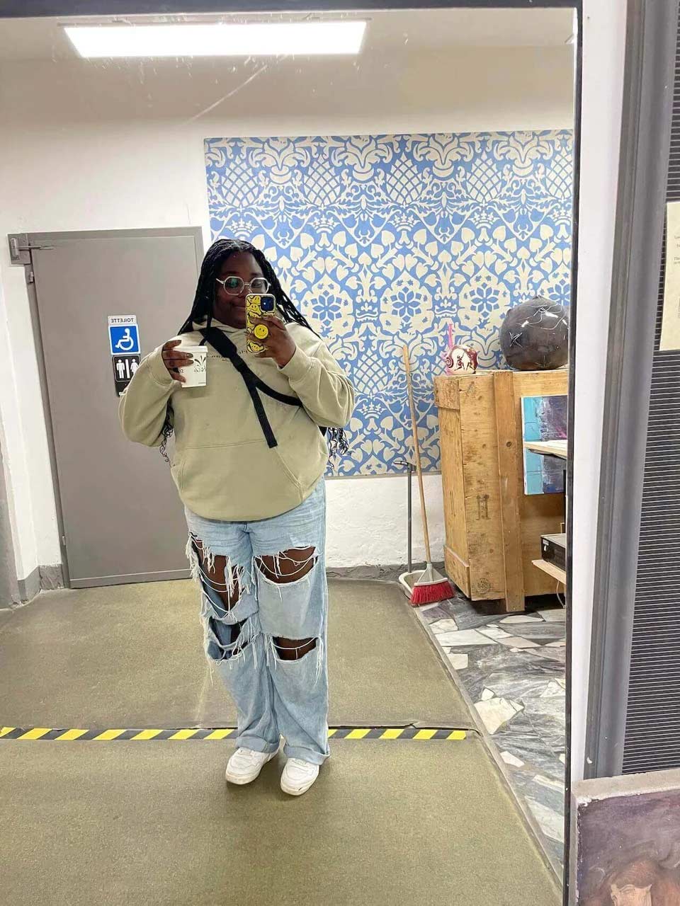 A student in hoodie and jeans holding a cup of coffee and taking a selfie in the mirror at an art studio