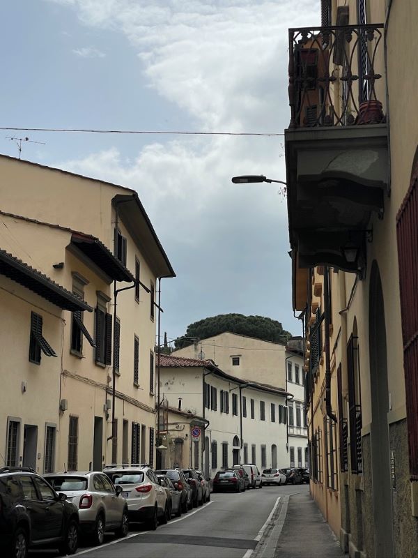 A city street hemmed in by light-colored buildings on either side and a line of cars parked along the lefthand side