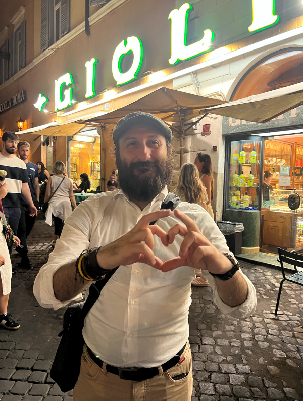 A person wearing a white shirt and flat cap standing on a cobblestone city street at night and making a heart sign with their fingers while smiling at the camera
