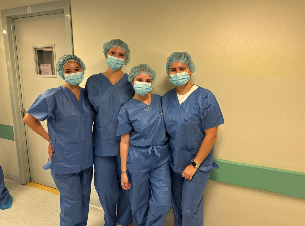 Four study abroad students wearing blue scrubs and disposable hair masks and face masks while standing in an indoor setting and smiling at the camera