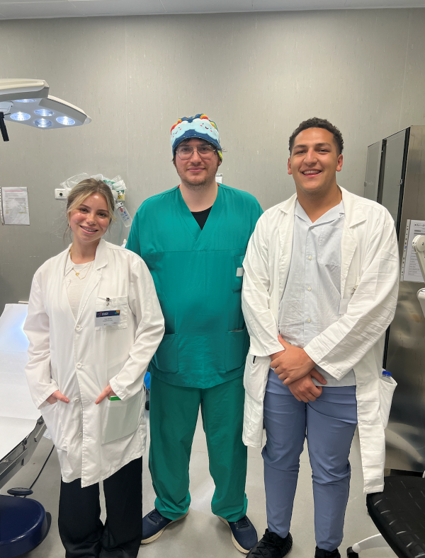 Two study abroad students wearing white medical coats, standing on either side of a medical professional wearing turquoise scrubs and a bandana, all three smiling at the camera
