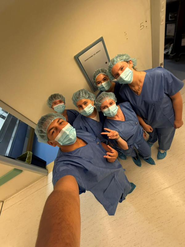 Six study abroad students taking a selfie in an indoor setting while wearing blue scrubs and disposable face and hair masks